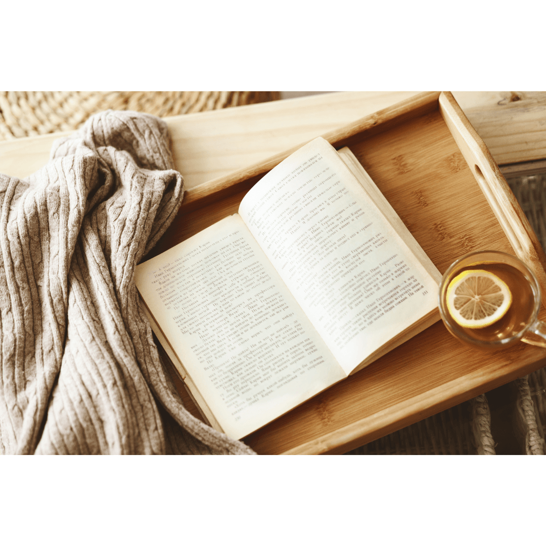 Overhead view of an open book on a wooden tray with a cup of lemon tea and a cozy wool throw blanket