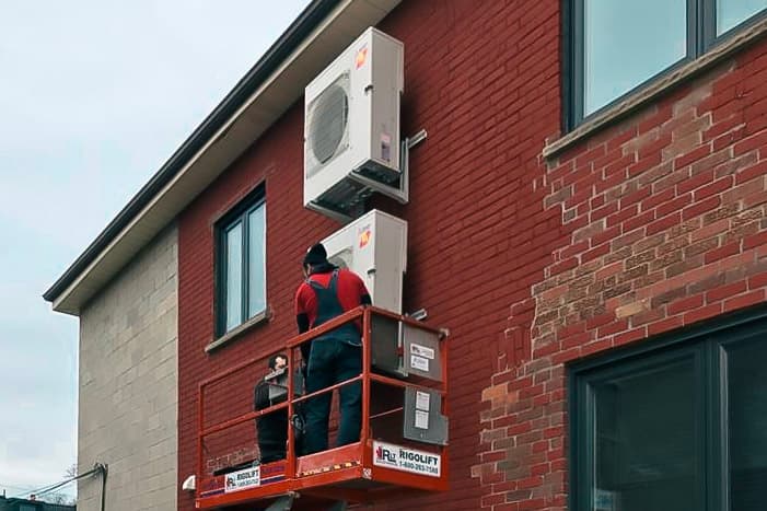 Two technicians are installing outdoor heat pump units on the brick wall of a residential building, using an aerial work platform for safe and precise work at height.