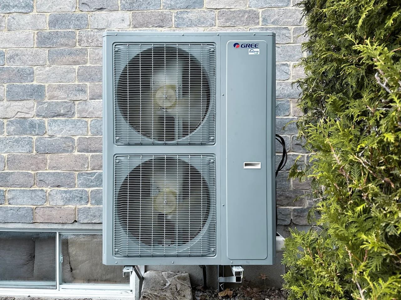 Gree outdoor heat pump unit with dual fans, installed on a stone wall in a residential setting, surrounded by greenery, offering an efficient solution for heating and cooling with a modern and robust design.