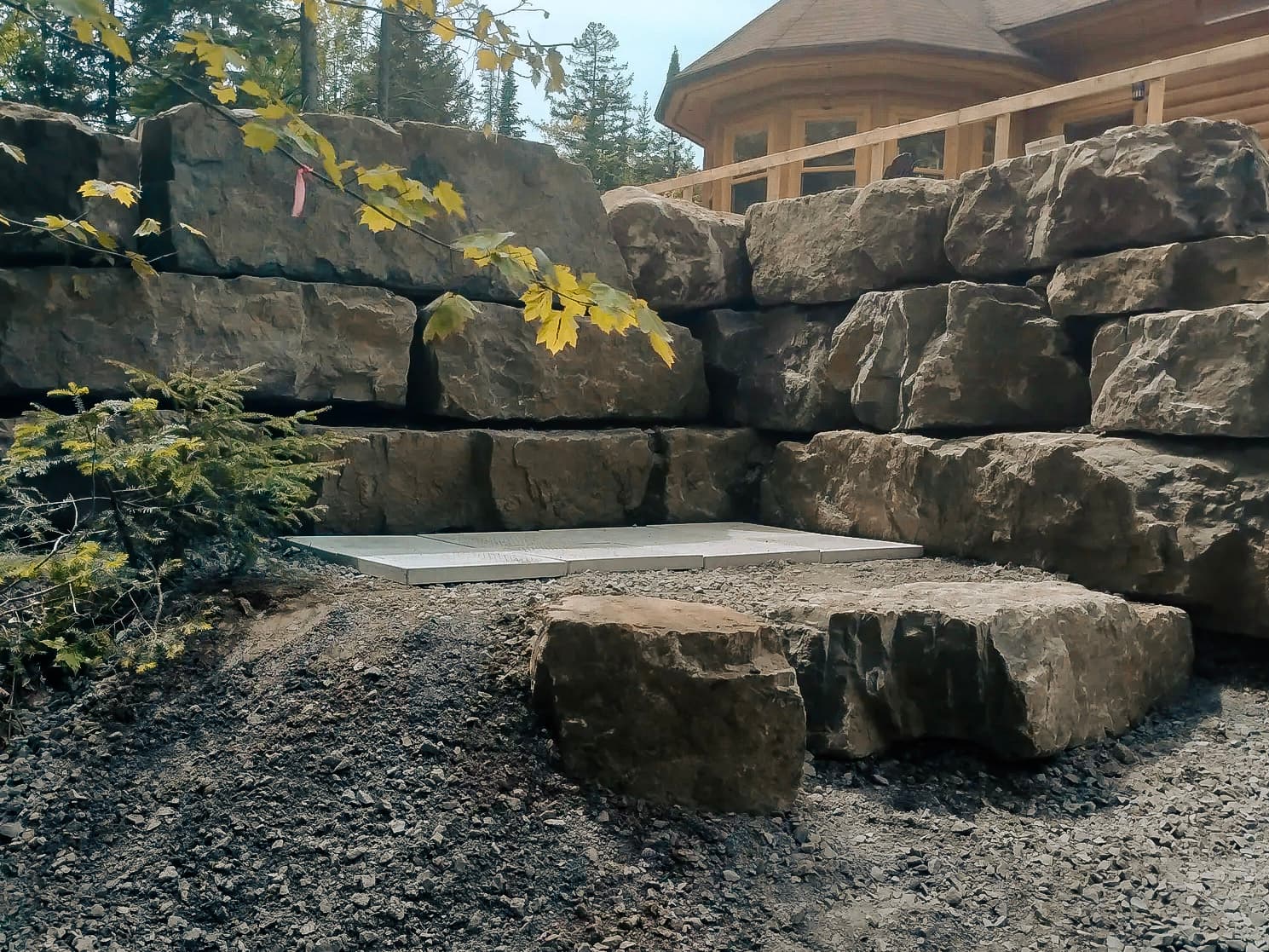 Massive stone rubble foundations forming a sturdy retaining wall, with a tree and foliage in the foreground, and a wooden house with a visible terrace in the background.