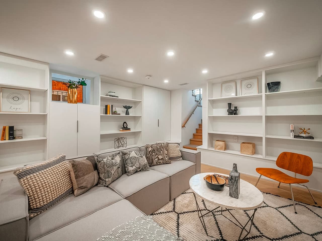 Finished basement with a modern living room featuring a modular sofa, built-in bookshelves, a marble coffee table, a wooden chair, and minimalist decor.