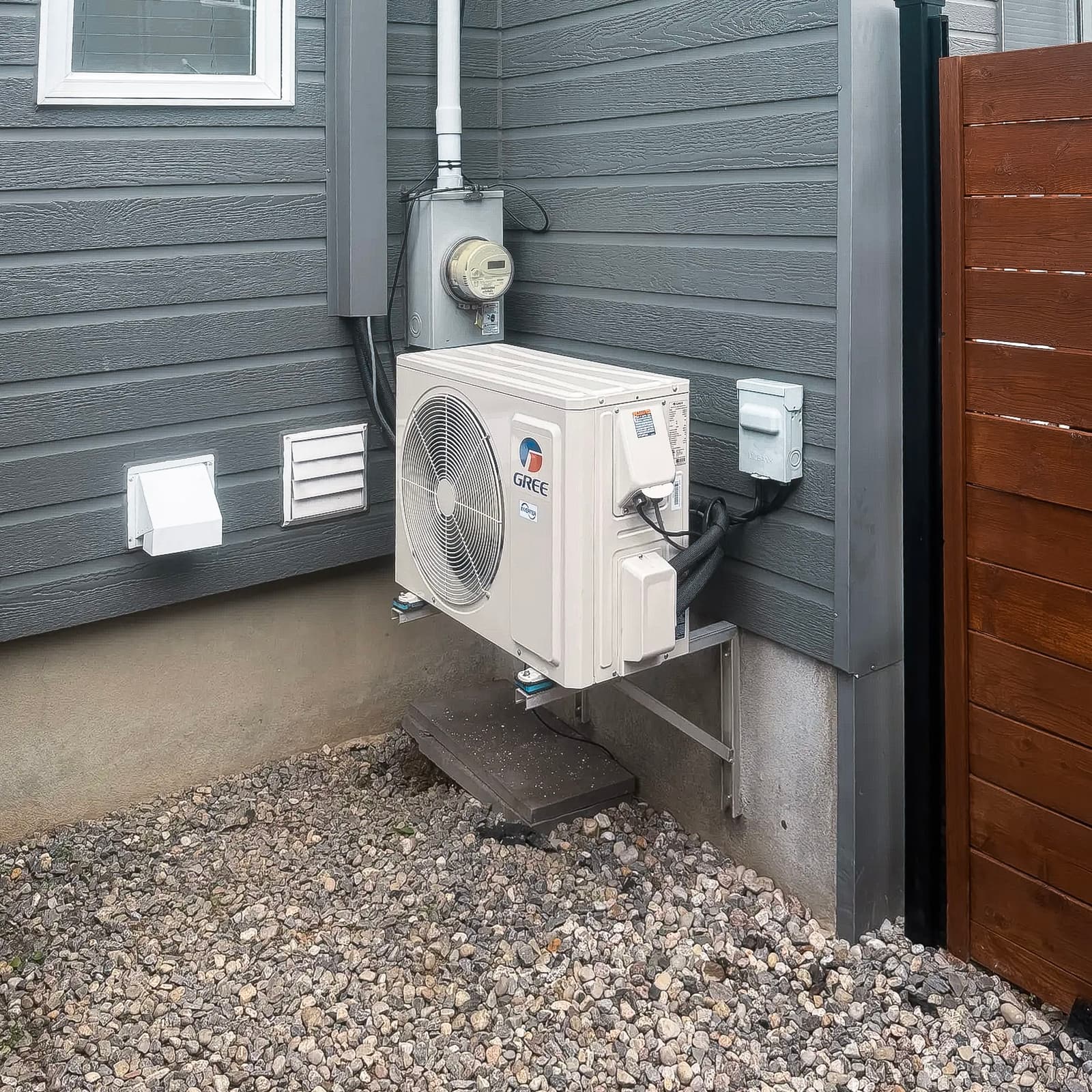 Outdoor unit of a Gree air conditioner installed on an exterior wall with gray siding and a gravel floor, next to a window and a wooden fence.<br>