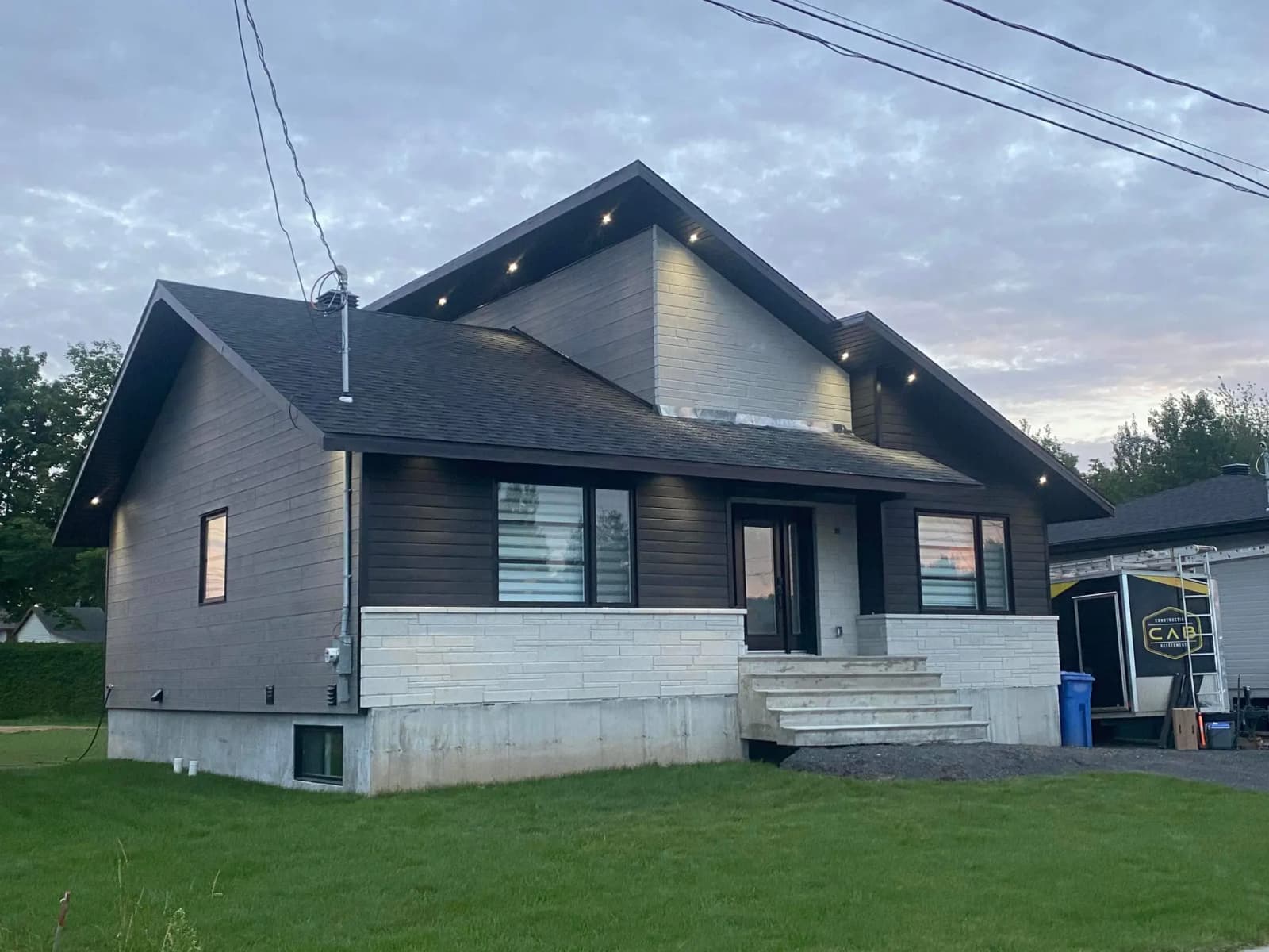A modern house with dark wood siding and white stone on the lower facade. It features a gabled roof with integrated lights under the eaves, a concrete staircase leading to the main entrance, and a visible garage on the right. The lawn is well-maintained, and the sky is slightly overcast at dusk.