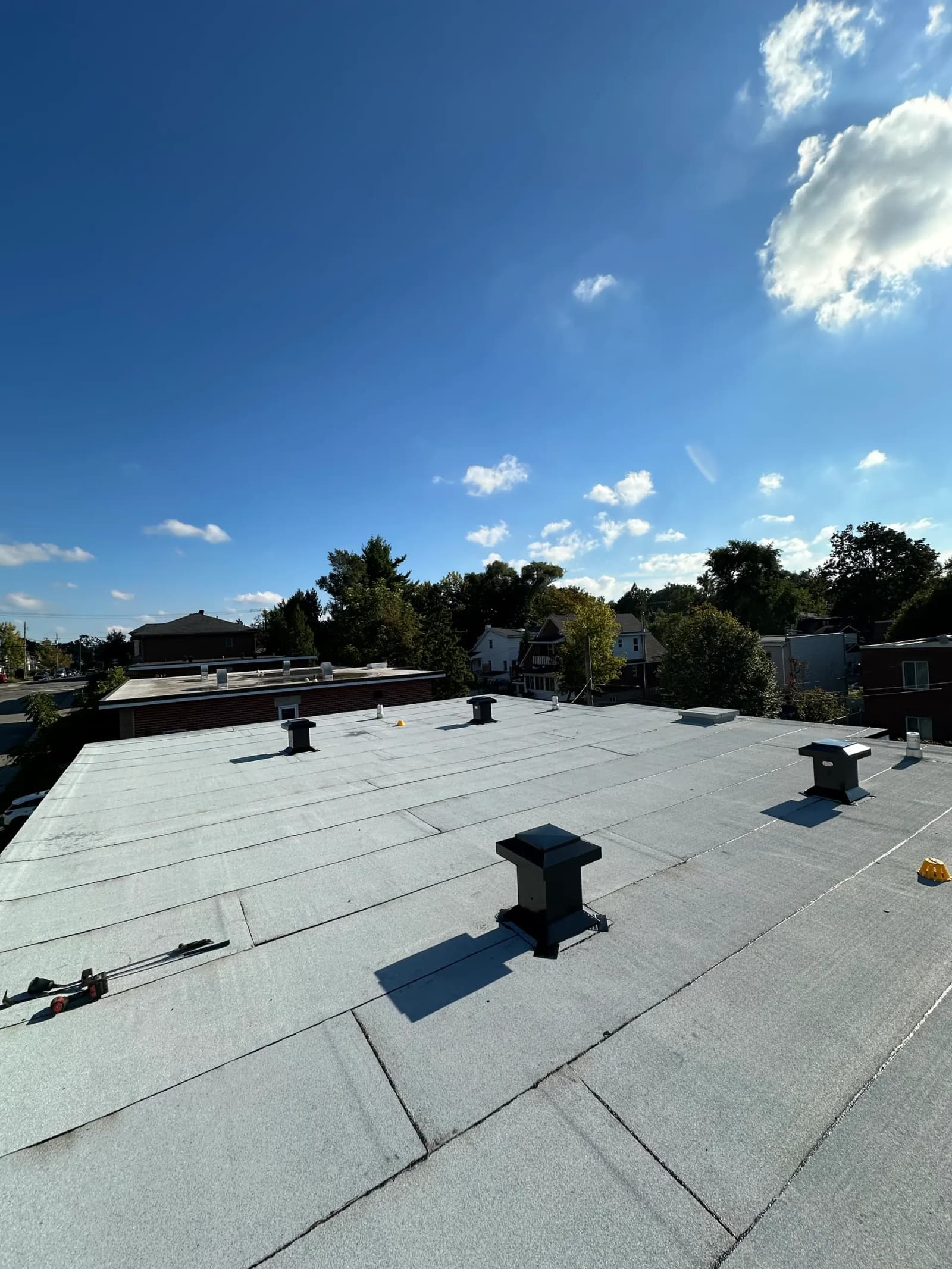 Flat roof covered with a gray membrane with several black ventilation ducts, overlooking a green residential neighborhood under a clear blue sky.