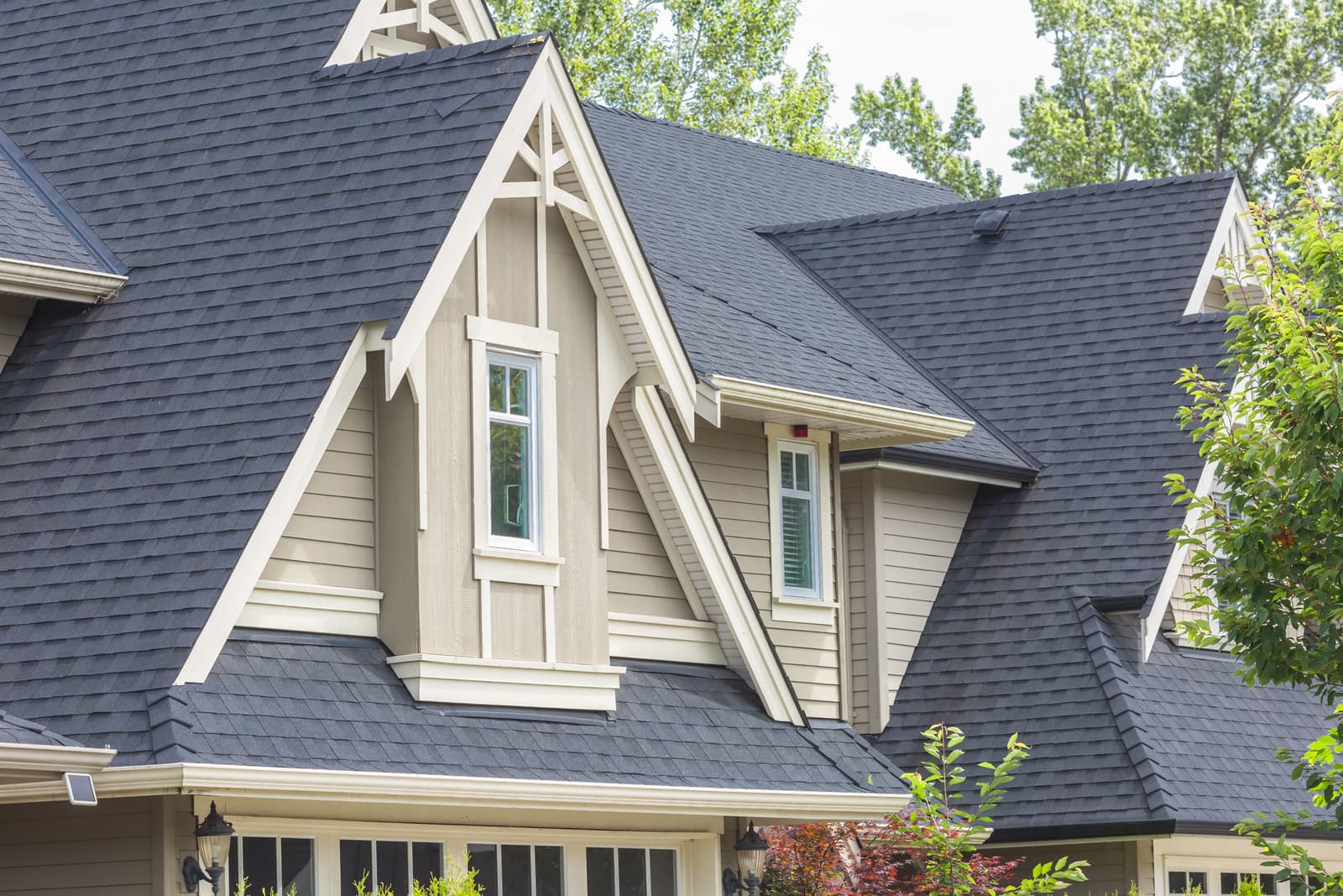 Close-up view of a house with a black asphalt shingle roof and beige exterior walls. The house features interesting architectural details, including decorative gables and sash windows.