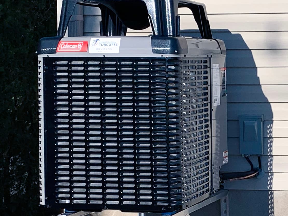 Outdoor air conditioning unit mounted on a metal wall bracket outside a house with beige vinyl siding.