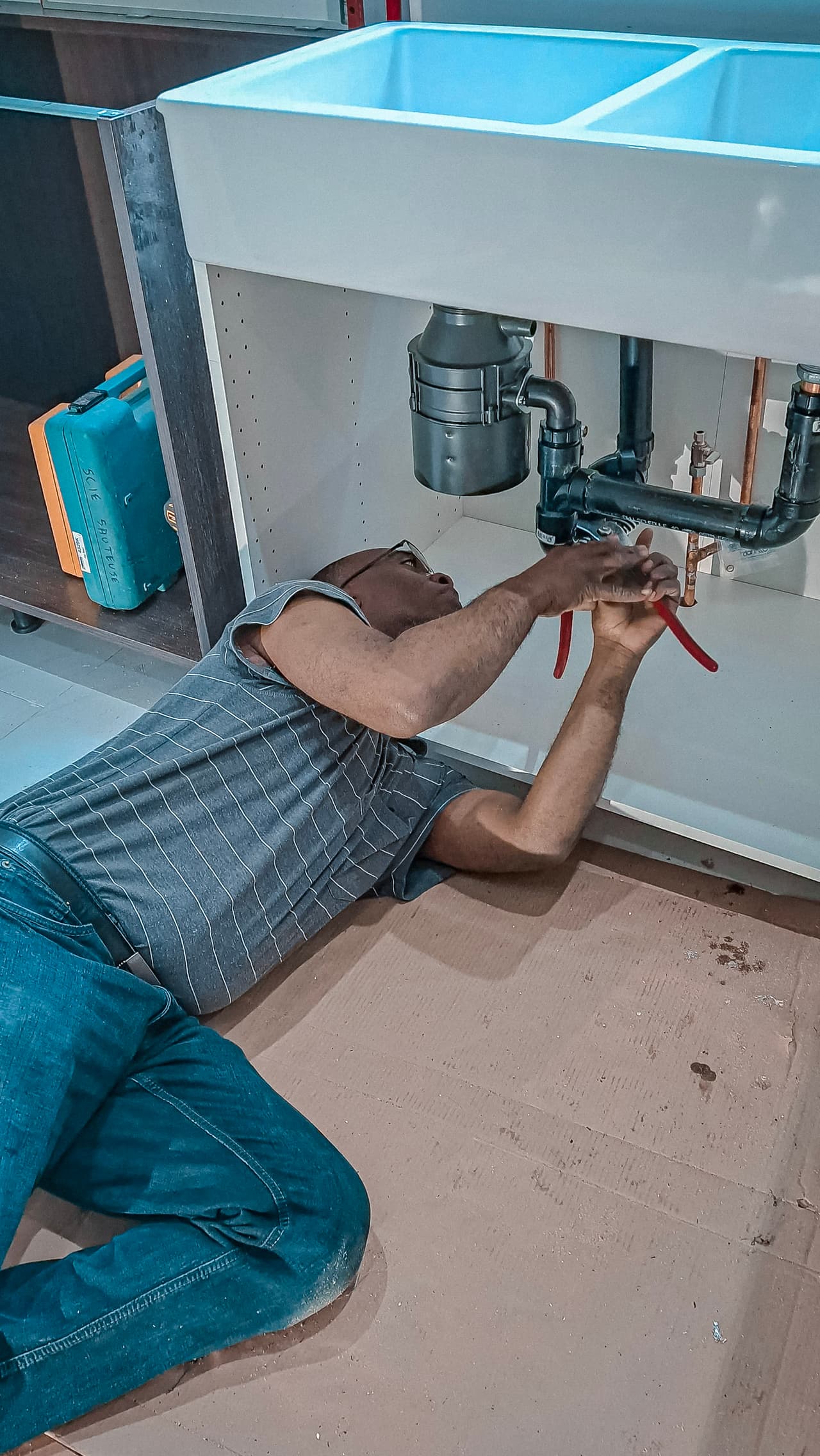 A plumber lying on the floor uses pliers to fix pipes under a kitchen sink.