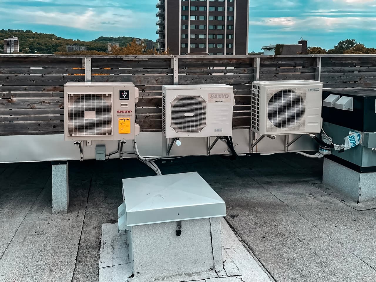 Outdoor air conditioning units installed on a flat rooftop with a view of residential buildings and green hills in the background.