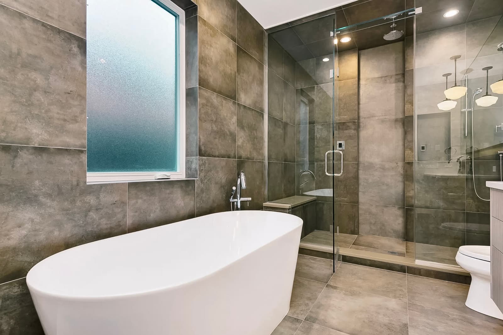 Modern bathroom with white freestanding bathtub, spacious glass shower, and stone-effect tiles on walls and floor. Frosted window for privacy and chrome fixtures.
