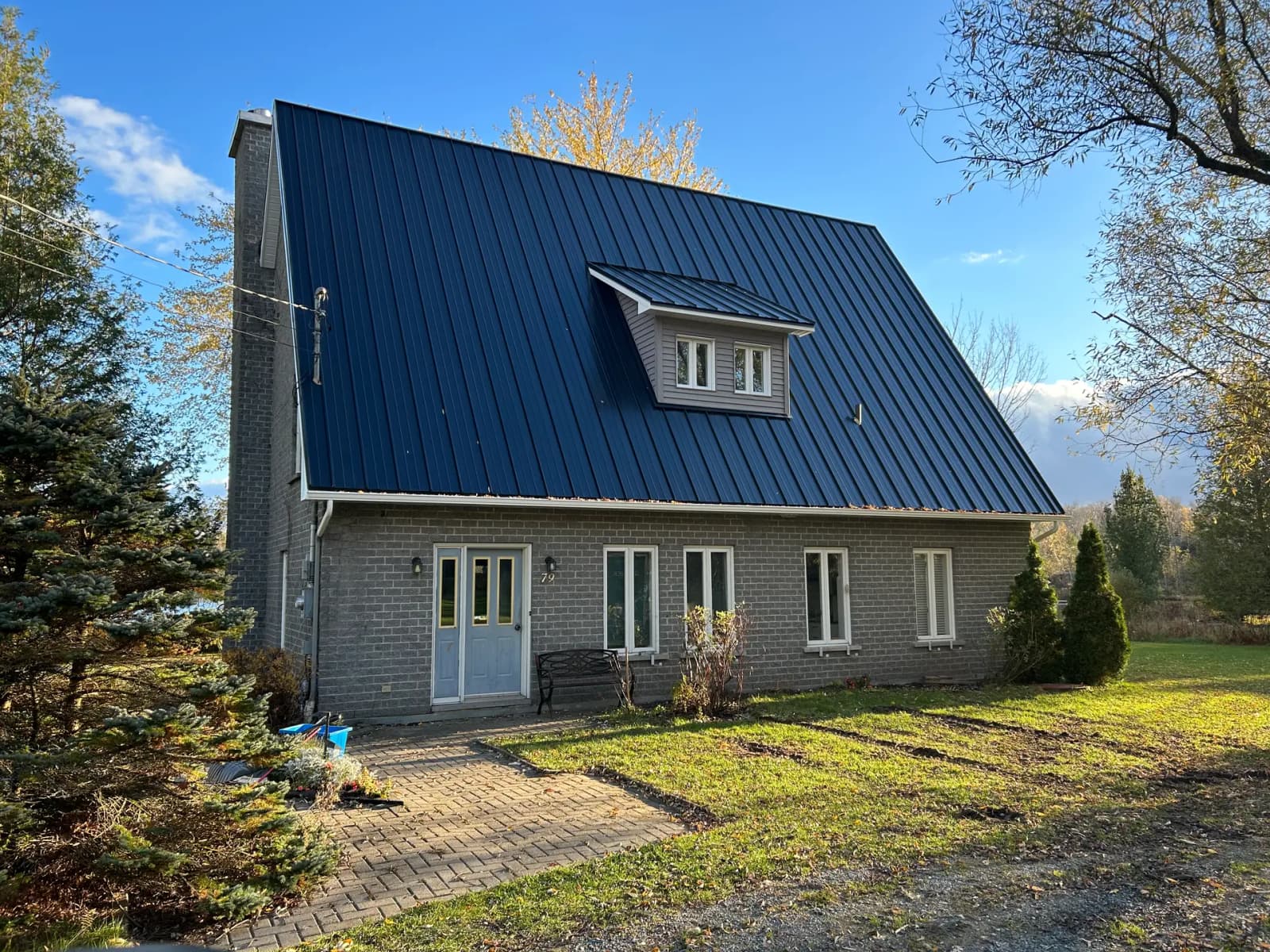 Brick house with a steeply sloped metal roof, featuring a dormer window in the center, surrounded by vegetation and trees in a sunny rural setting.