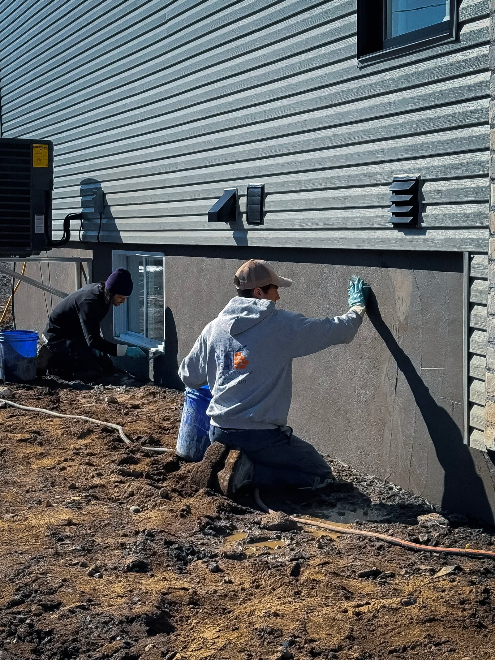Workers applying a coating on the exterior foundation of a vinyl-sided house.