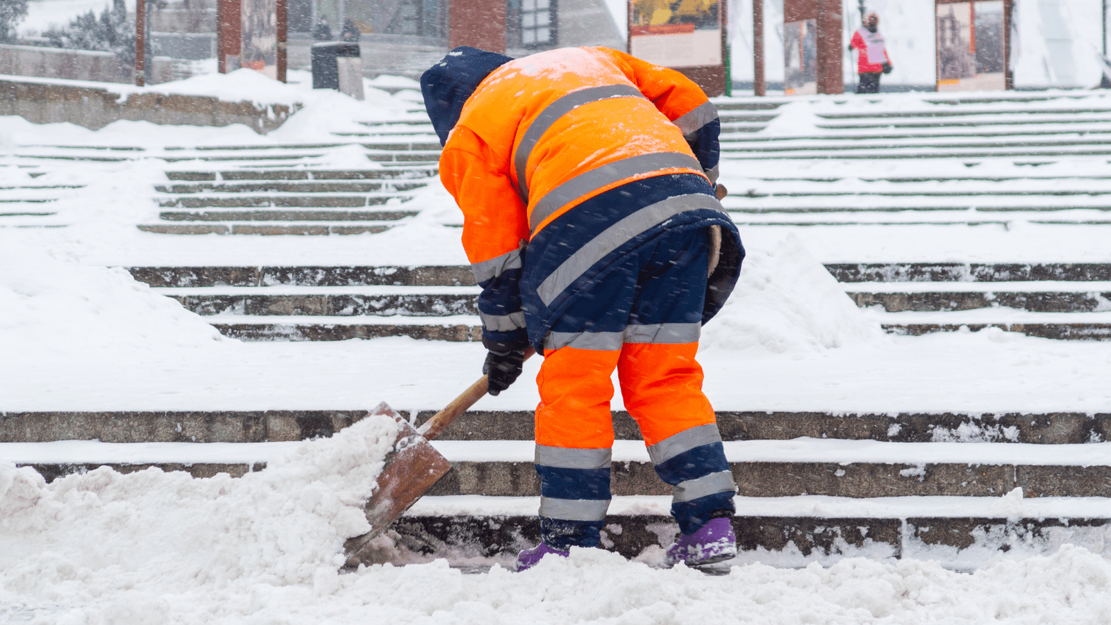 Worker in high-visibility clothing using a shovel to clear snow from stairs during winter conditions.