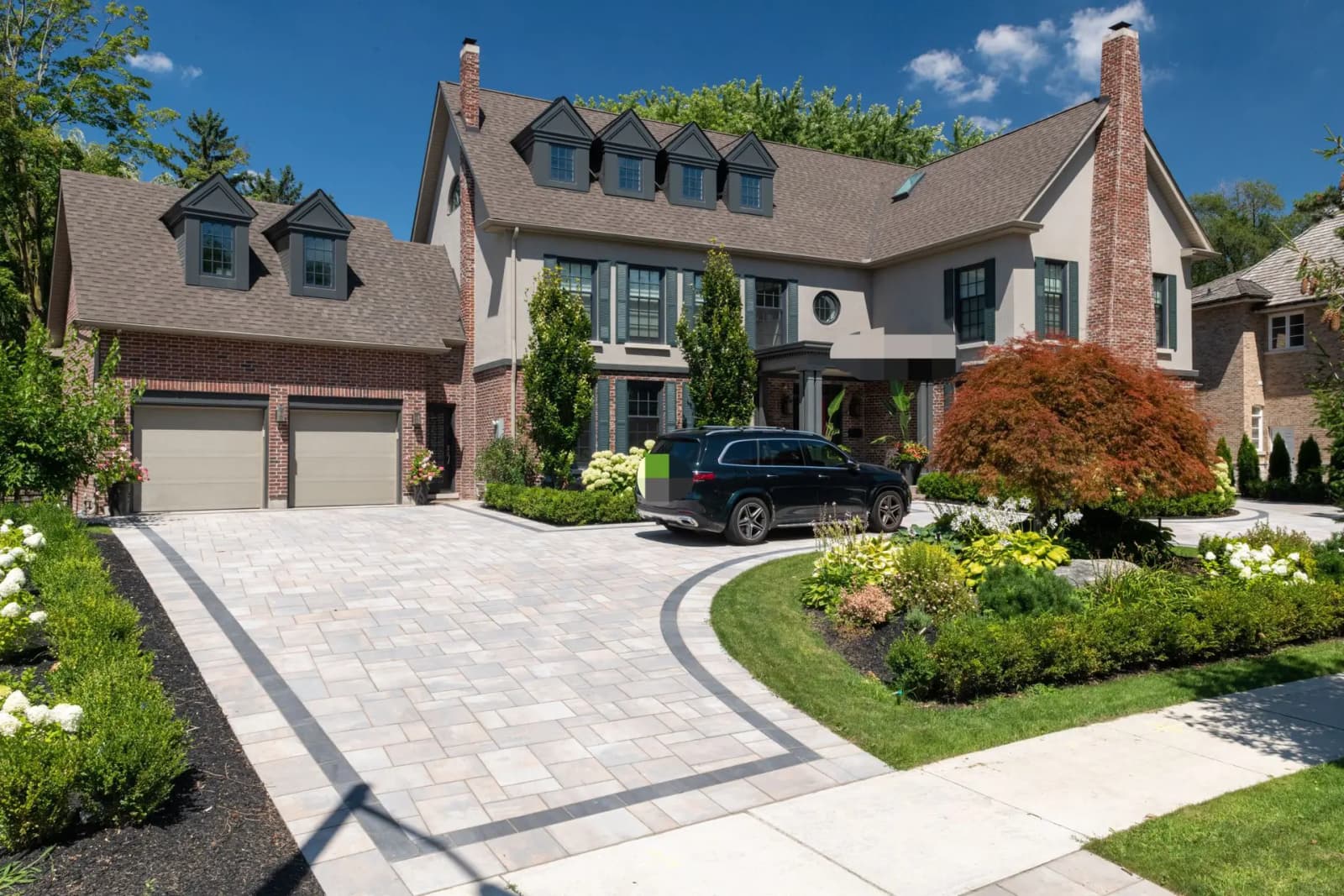 Large traditional-style house with a spacious paved entrance, a well-maintained landscaped garden, and a car parked in front of the garage under a sunny sky.