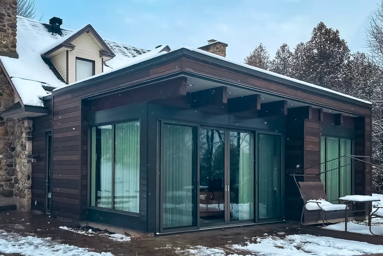 Modern house extension with dark wood siding, large sliding glass doors, flat roof, and snow-covered patio.