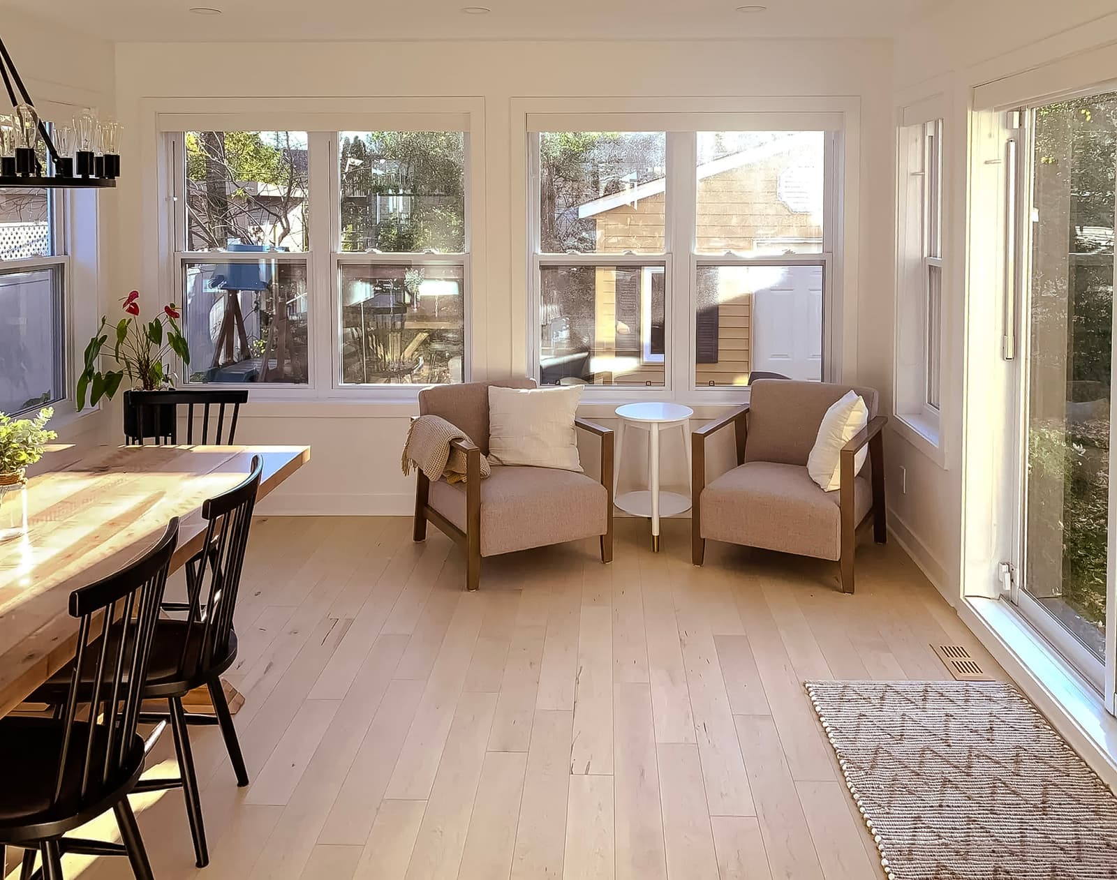 Bright dining and sitting area with large windows, light wood flooring, solid wood table, and black chairs. Two beige armchairs with cushions and a white side table.