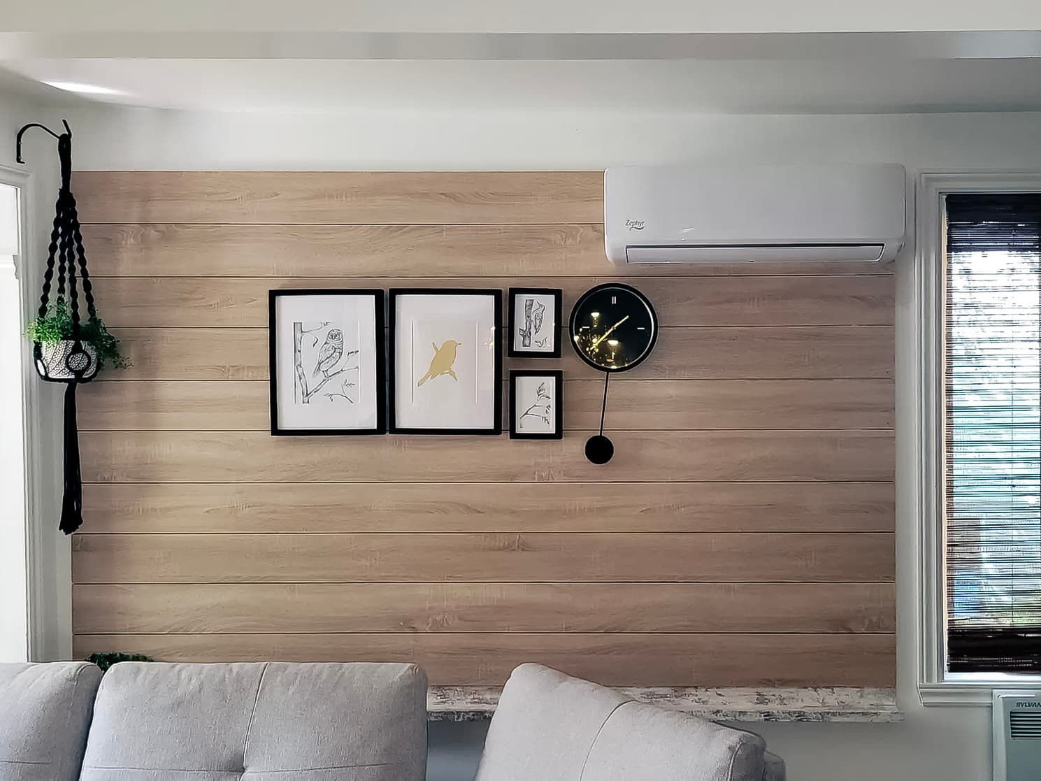 Light wood wall with wall-mounted air conditioner, decorative frame gallery, black designer clock, and hanging plant in a modern living room.