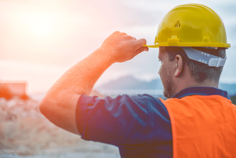 Construction worker wearing a yellow hard hat and orange safety vest, looking out over a job site at sunrise, symbolizing continuity and vigilance despite the residential sector strike.