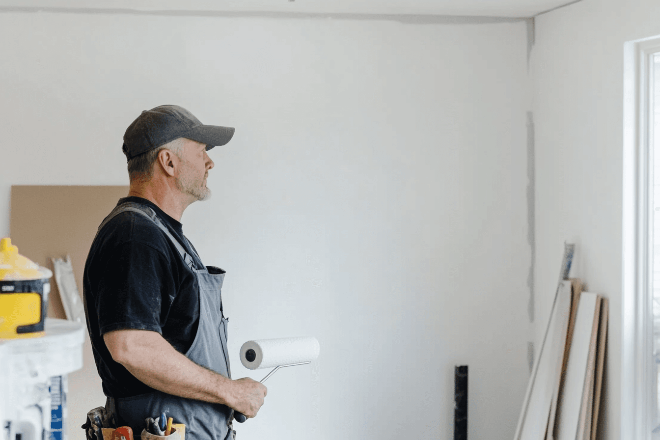 Building painter wearing overalls and a cap, holding a paint roller in a room under renovation.