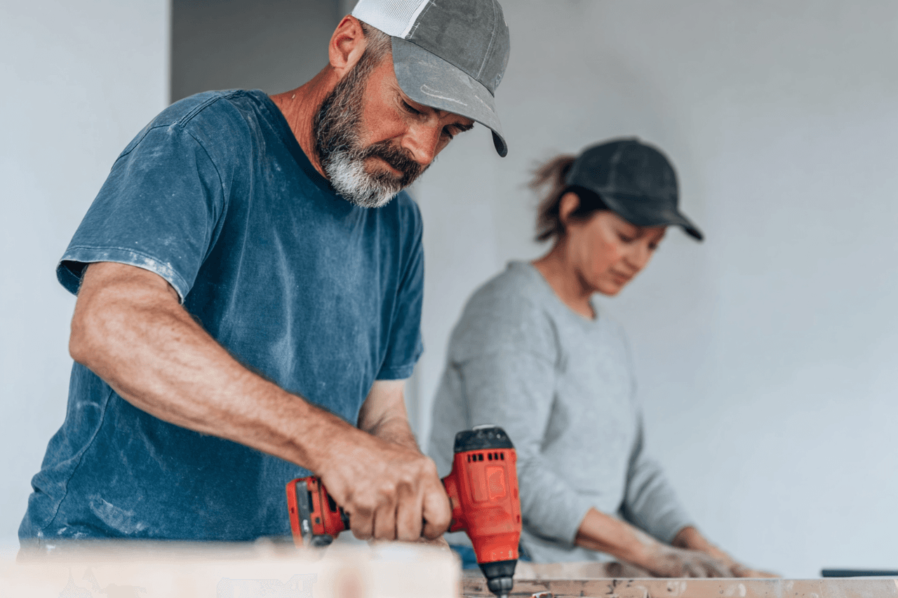 Interior renovation contractor using a drill with a coworker on a residential job site, surrounded by drywall, wood boards, construction dust and tools for carpentry and installation work
