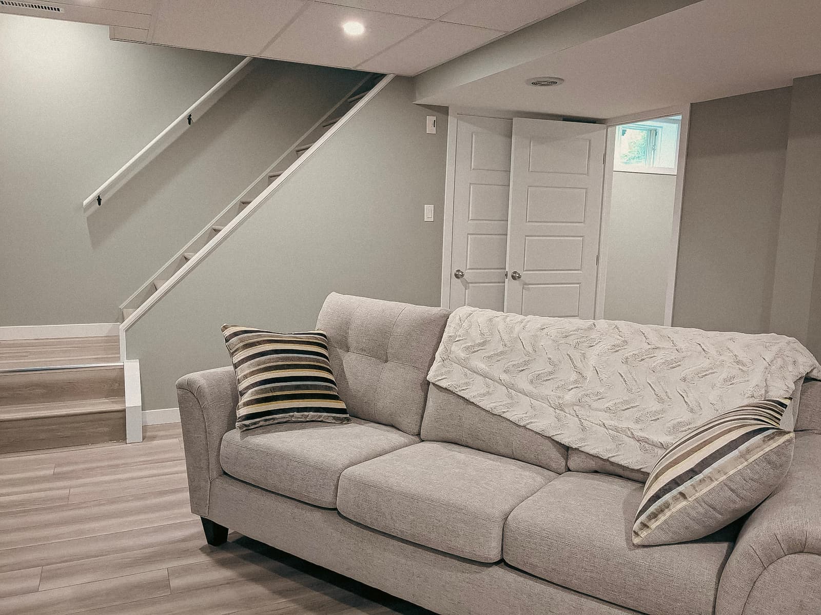 Finished basement with a gray sofa, wooden stairs, and a neutral wall with a white door.