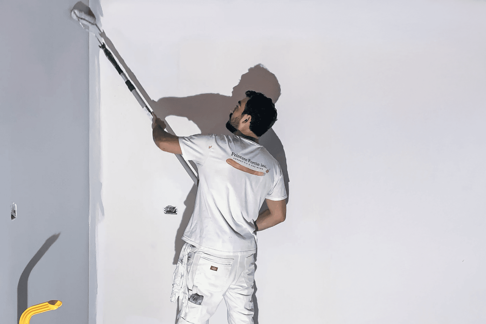 Building painter applying a coat of white paint on a wall using a telescopic roller, wearing white work attire.