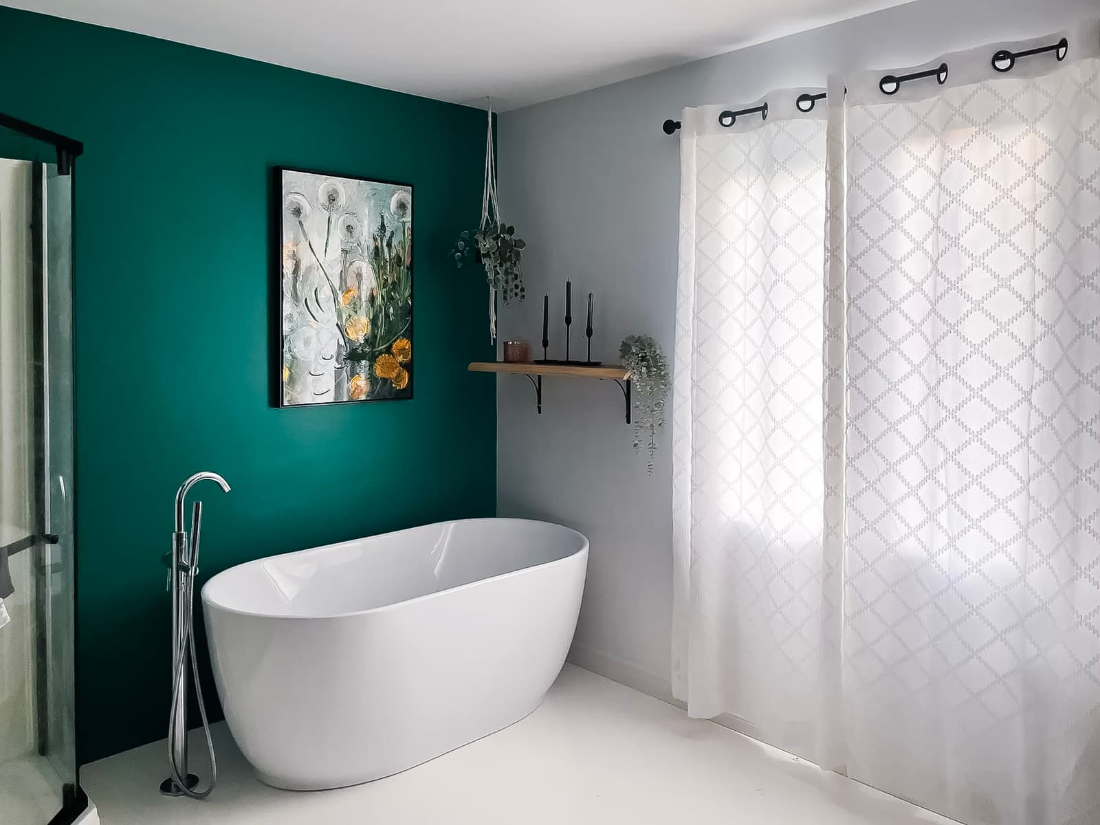Modern bathroom with emerald green wall, white freestanding bathtub, floral painting, wooden shelf with hanging plants, and sheer white curtains.