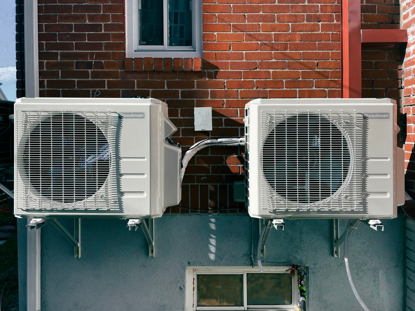 Two Goodman air conditioning units mounted on a brick wall with metal brackets.