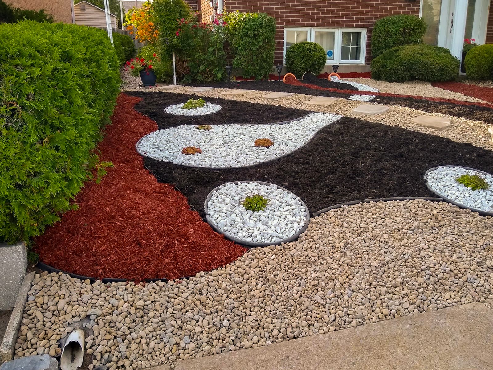 Yin-yang inspired landscaping with colored mulch, decorative stones, and succulent plants in front of a brick house.