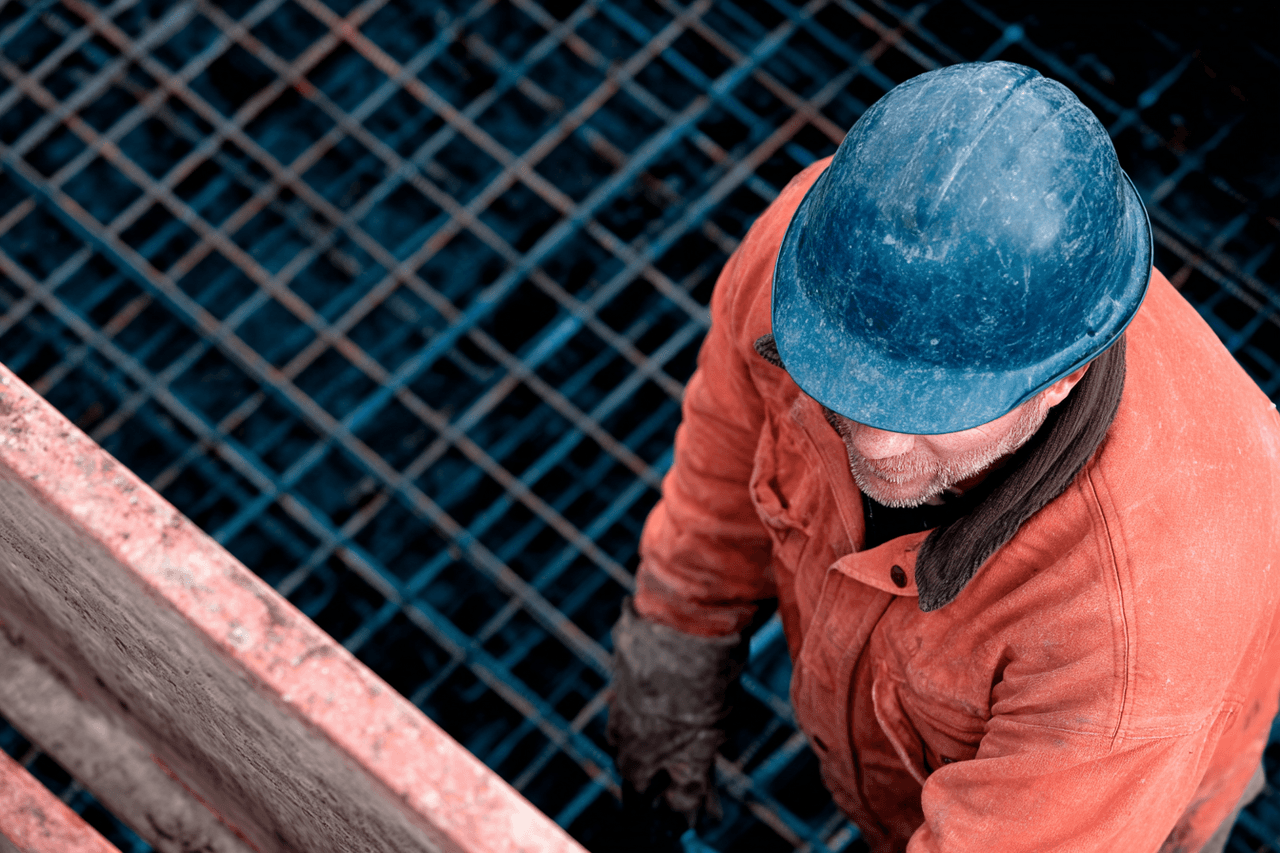 Construction worker wearing a blue helmet and orange jacket, standing on a steel reinforcement grid for concrete foundation.
