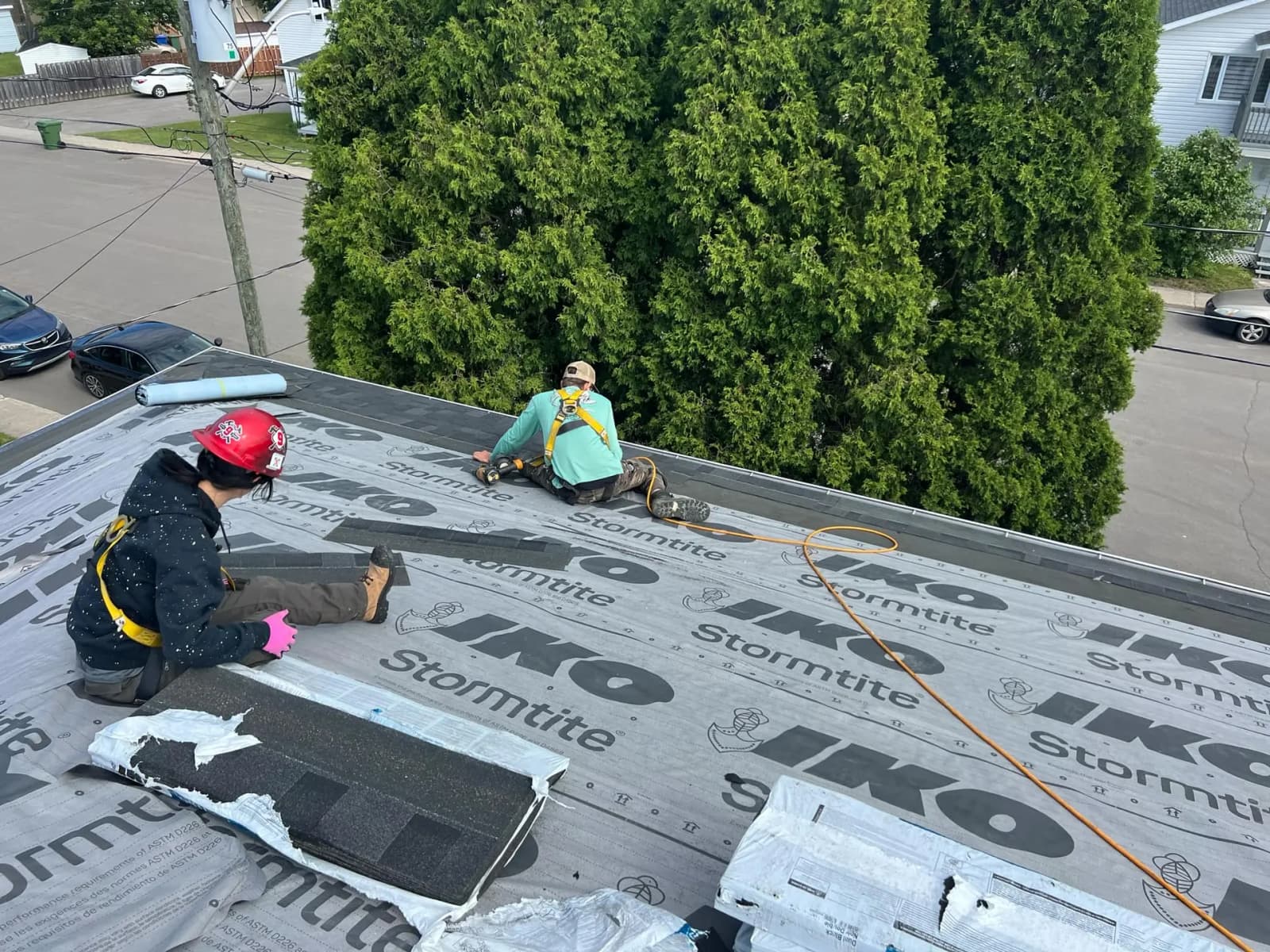 Two roofers are installing shingles on a roof with an IKO Stormtite underlayment. They are wearing safety equipment and working near a large hedge of green trees, with a street and cars visible in the background.