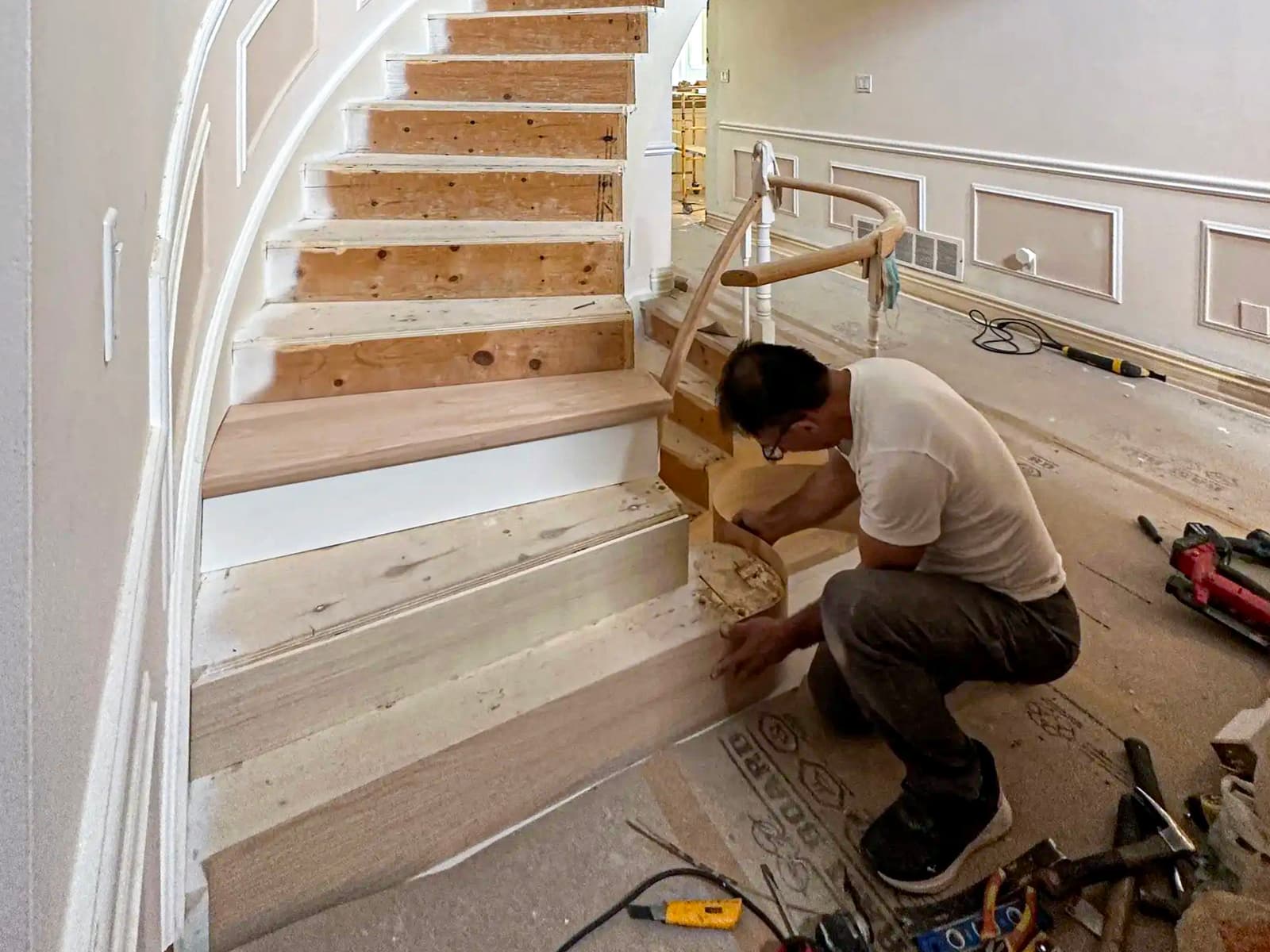 Curved wooden staircase under renovation, with a craftsman working on the finishing touches in a classic interior with wall moldings.
