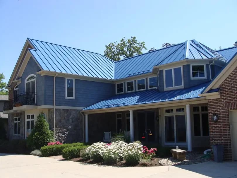 Modern two-story house with a blue metal gable roof and multiple slopes, combined with blue siding and a stone facade. The metal roof provides an elegant and durable finish