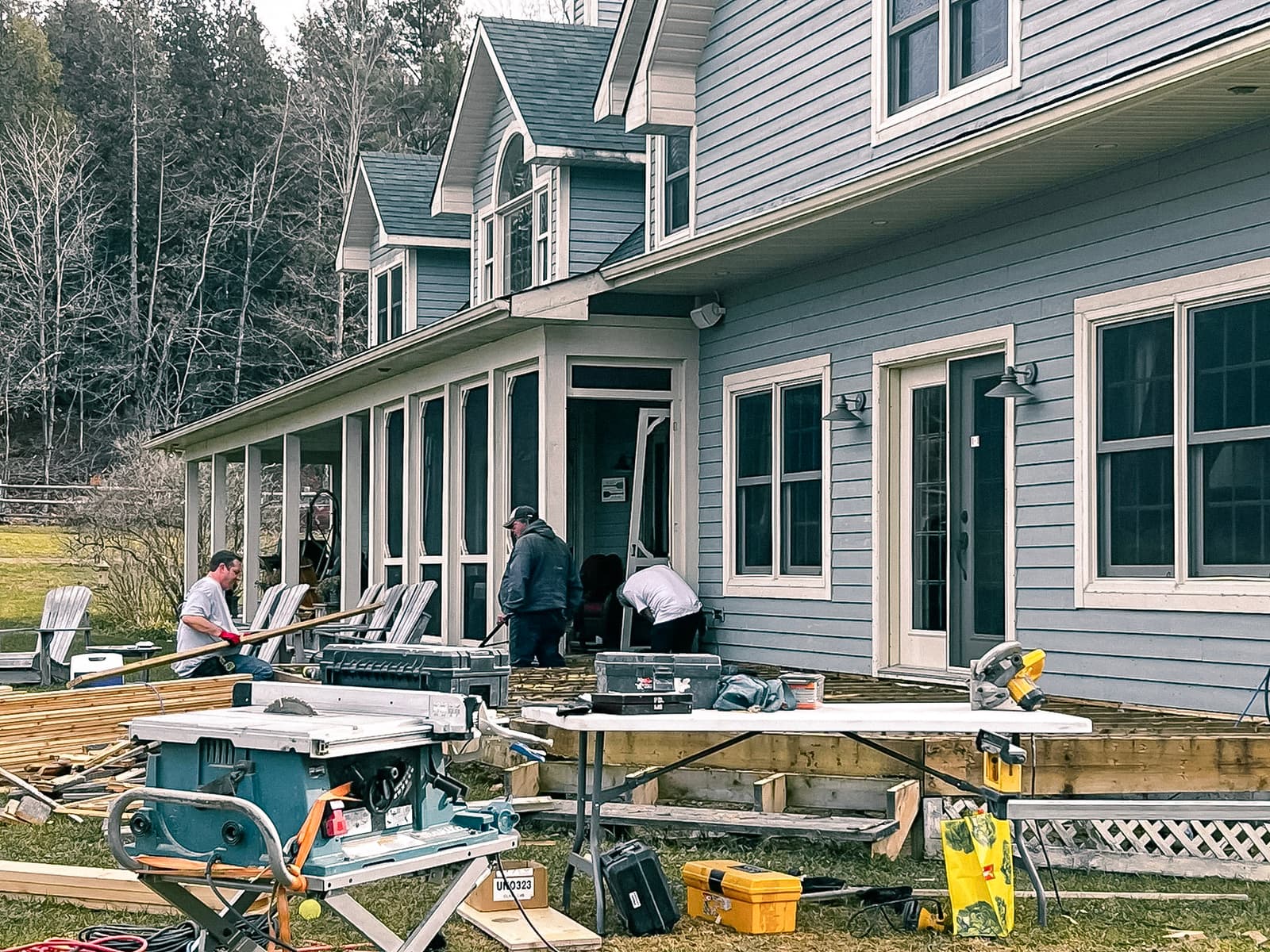 Workers renovating the wooden deck of a blue house with tools and construction equipment.