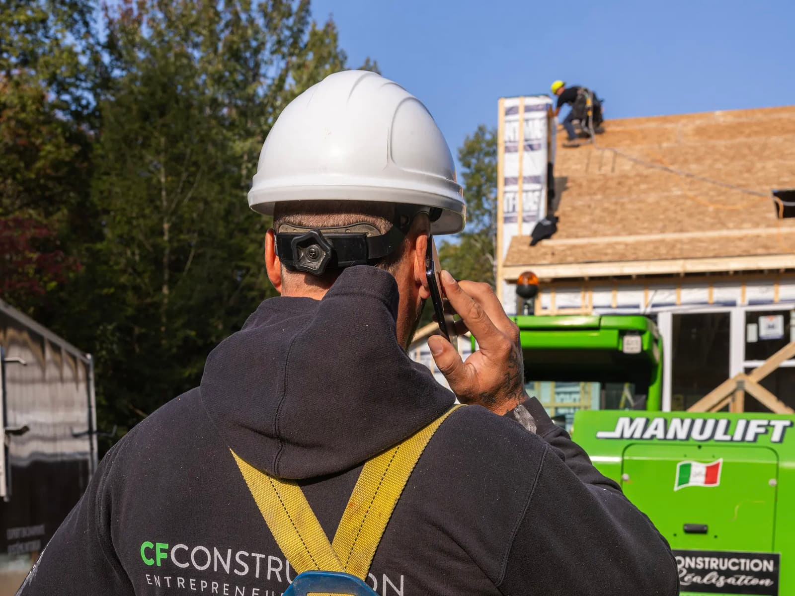 Construction worker wearing a safety harness and helmet, talking on the phone at a job site with roofing work in the background.