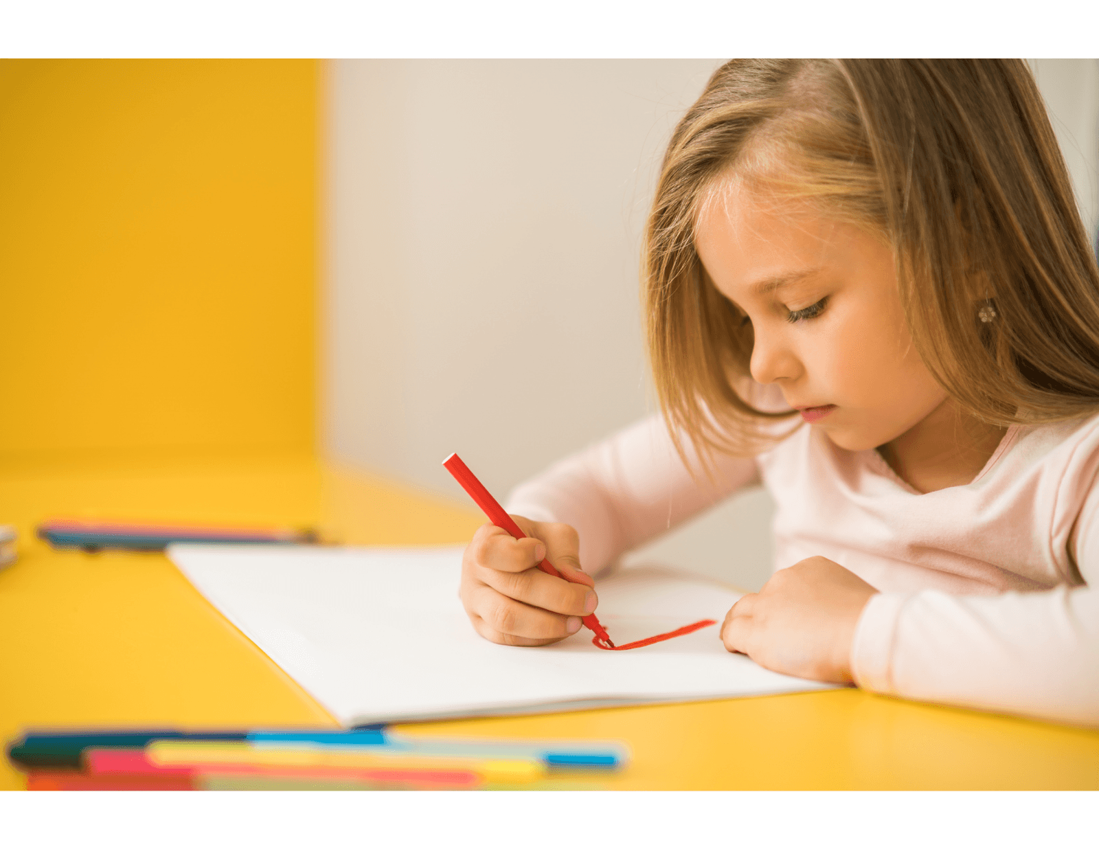 &nbsp;Little girl drawing with a red pencil on a sheet of paper, sitting at a yellow table.