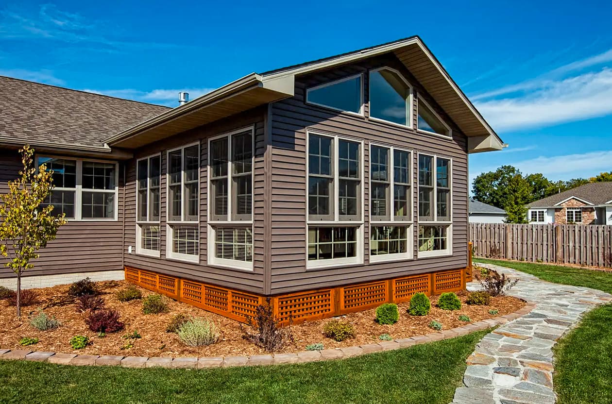 Home extension with a large glass sunroom, wood siding, landscaping with mulch and a stone pathway, blue sky in the background.