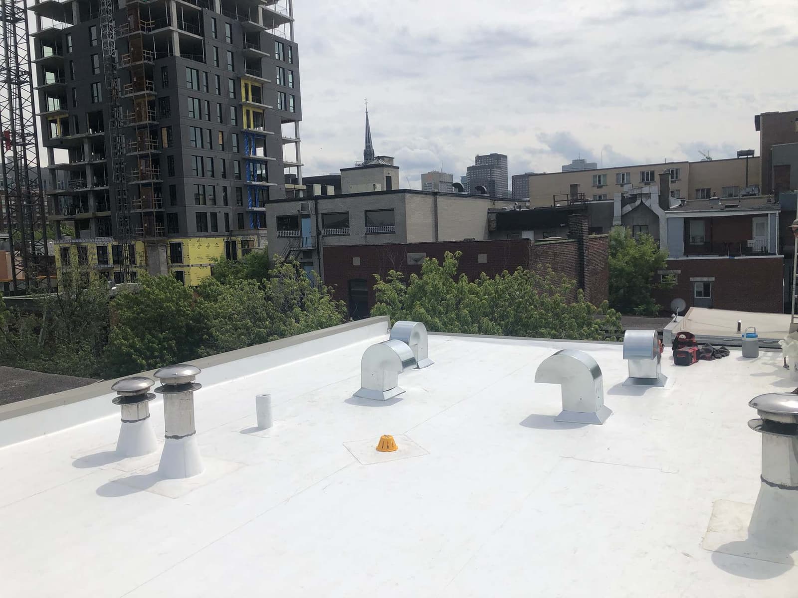 Flat roof covered with white membrane, equipped with several ventilation chimneys and ducts, overlooking an urban neighborhood under development with residential and commercial buildings in the background.&nbsp;