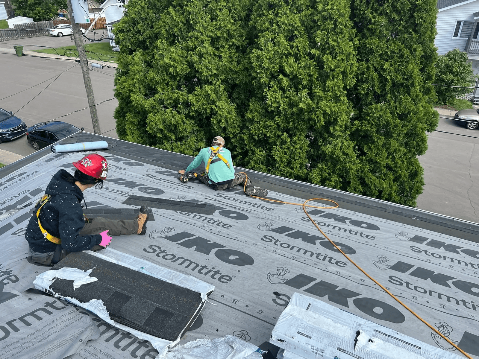 Two roofers are installing shingles on a roof with an IKO Stormtite underlayment. They are wearing safety equipment and working near a large hedge of green trees, with a street and cars visible in the background. Select an Image