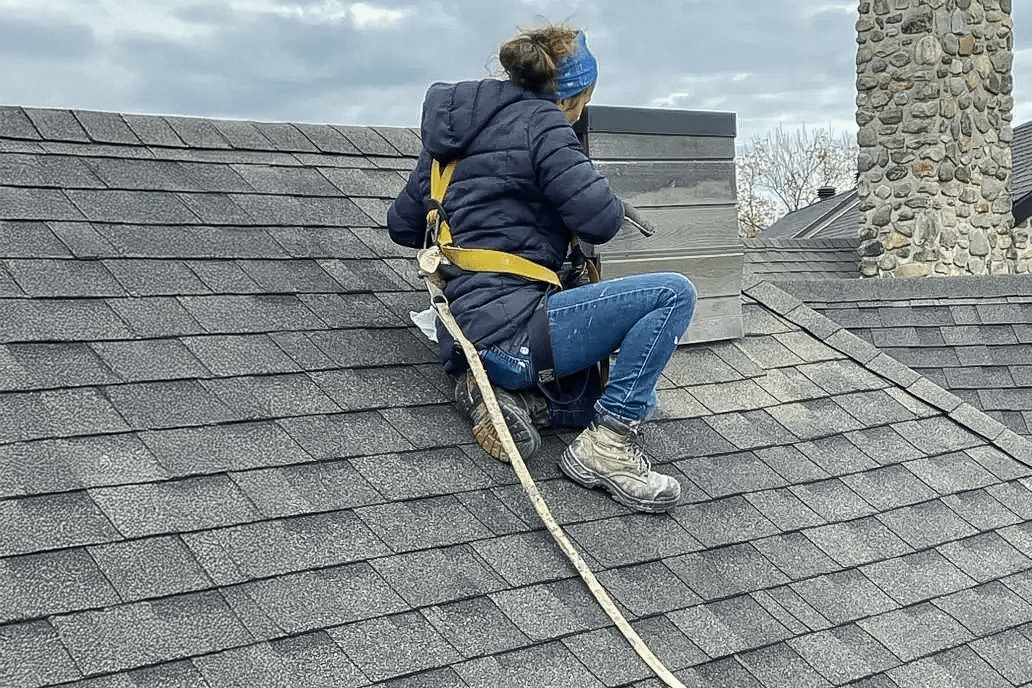 Worker in a safety harness performing work on an asphalt shingle roof, near a wooden-clad vent pipe.