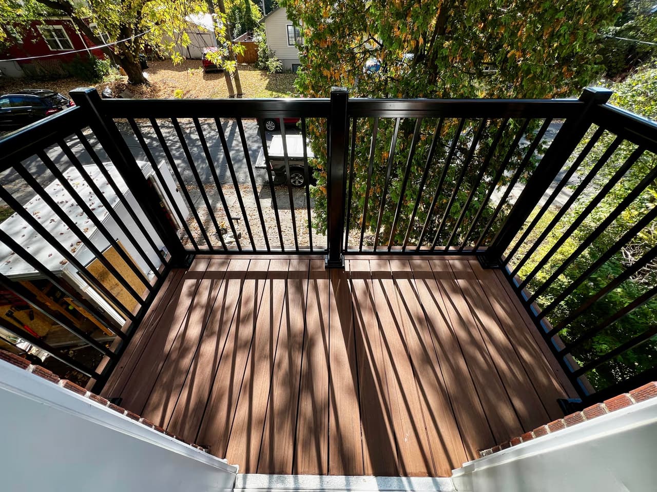 Outdoor balcony with brown composite wood flooring and black railing overlooking a tree-lined street.