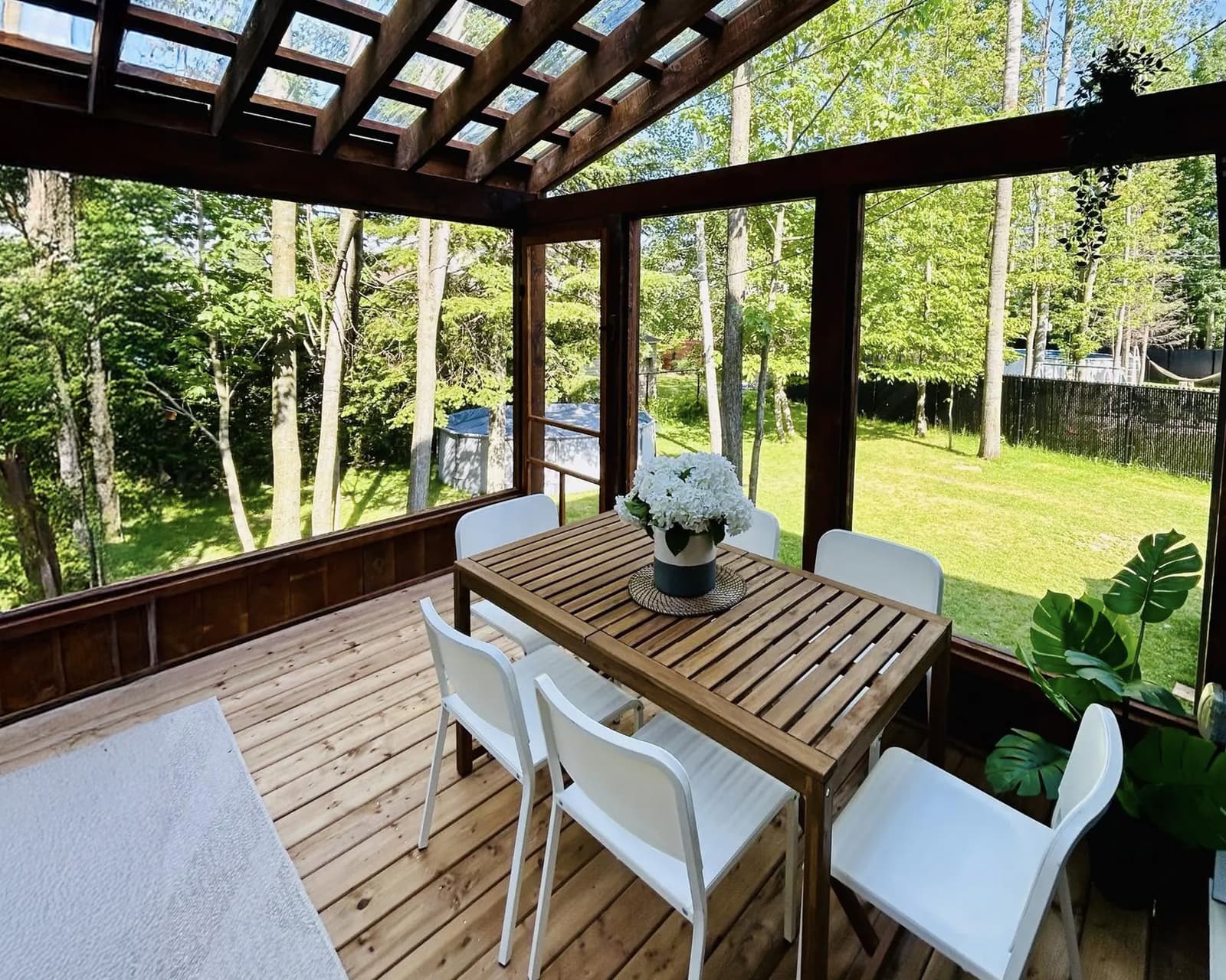 Wooden veranda with glass roof, wooden garden table, and white chairs, providing a bright and modern outdoor space surrounded by greenery.