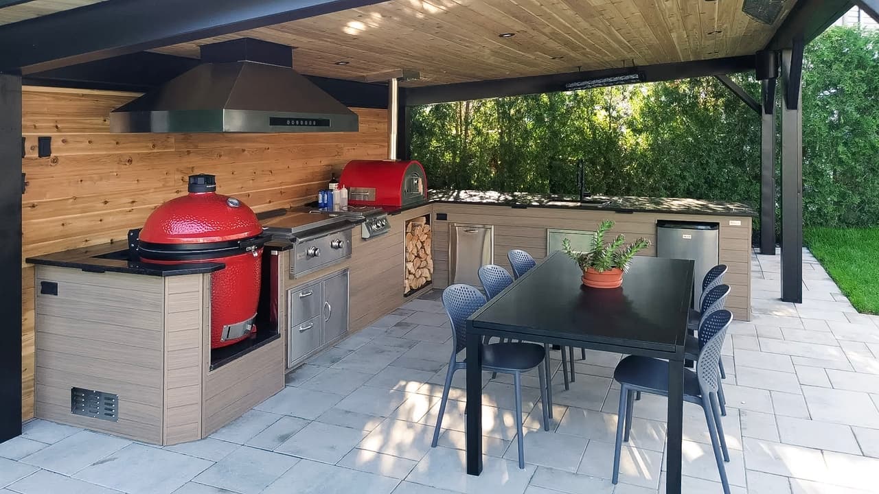 Modern outdoor kitchen with red ceramic barbecue, pizza oven, wood countertops, and covered dining area