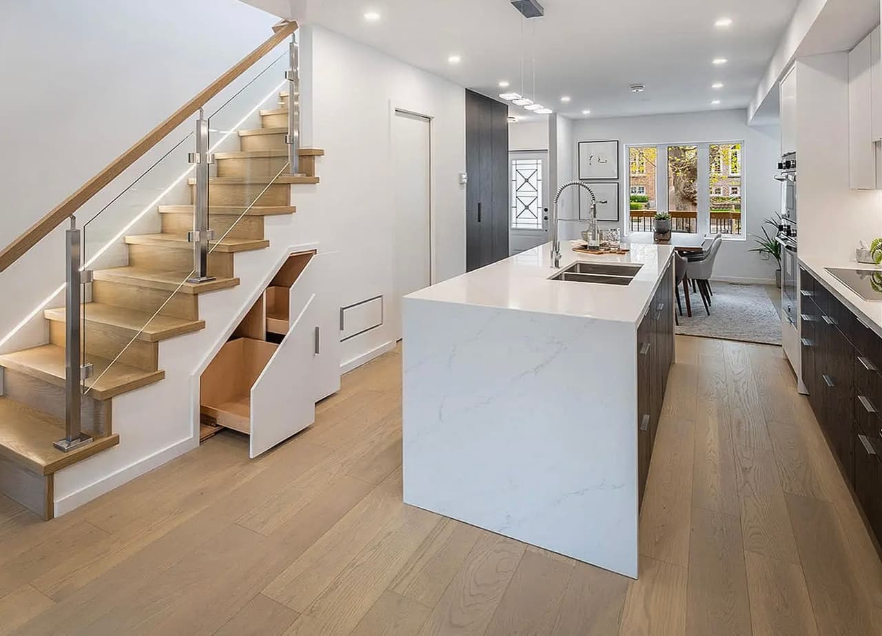Modern home interior with open kitchen, wooden staircase with built-in storage, and white marble island. Bright dining area with garden access.