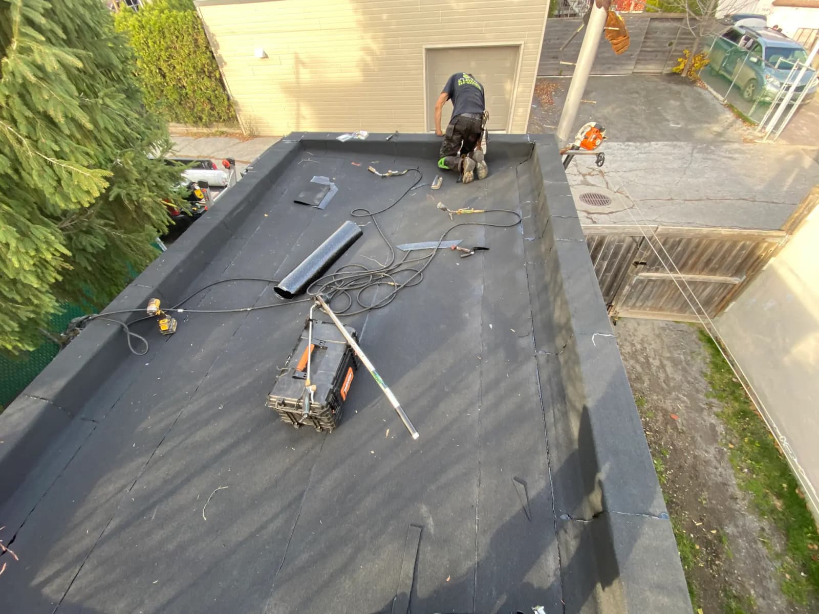Installation work of a new black membrane on a flat roof, with a worker actively laying it, surrounded by tools and equipment in a fenced residential yard.