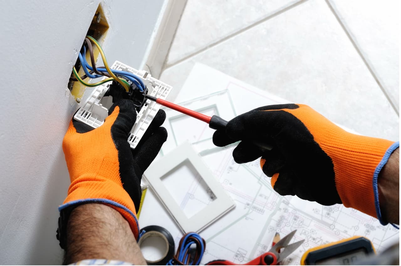 Electrician wearing orange and black gloves repairing or installing a wall outlet, using a screwdriver with visible electrical wires.