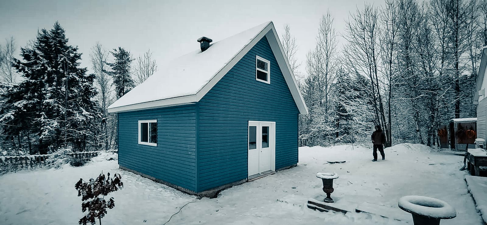 Small blue wooden house with a snow-covered roof, located in a winter forest. Peaceful setting with snow-covered trees and a person in the background.