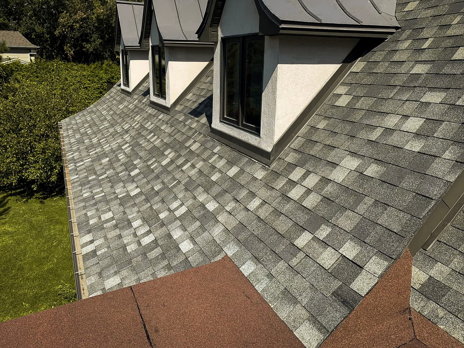 Residential roof with asphalt shingles and dormer windows overlooking a lush green garden.<br>
