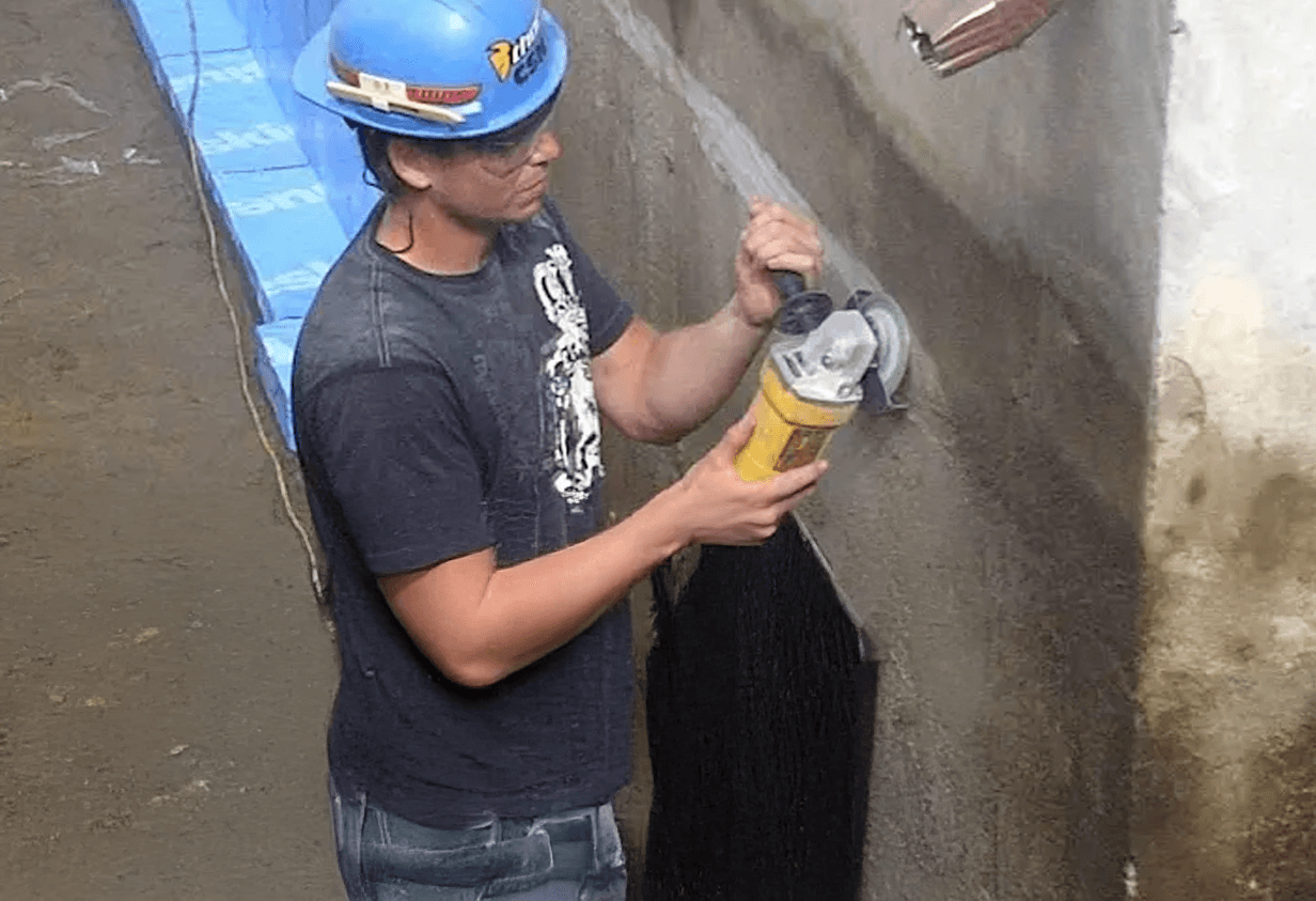 Worker with helmet grinding a concrete foundation in preparation for waterproofing application