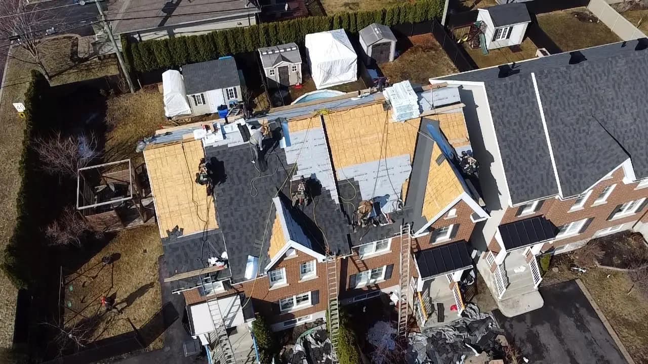 Asphalt shingle roof renovation work on a brick semi-detached house, with workers installing the new covering on the exposed framework, surrounded by several outbuildings and gardens in a residential yard.