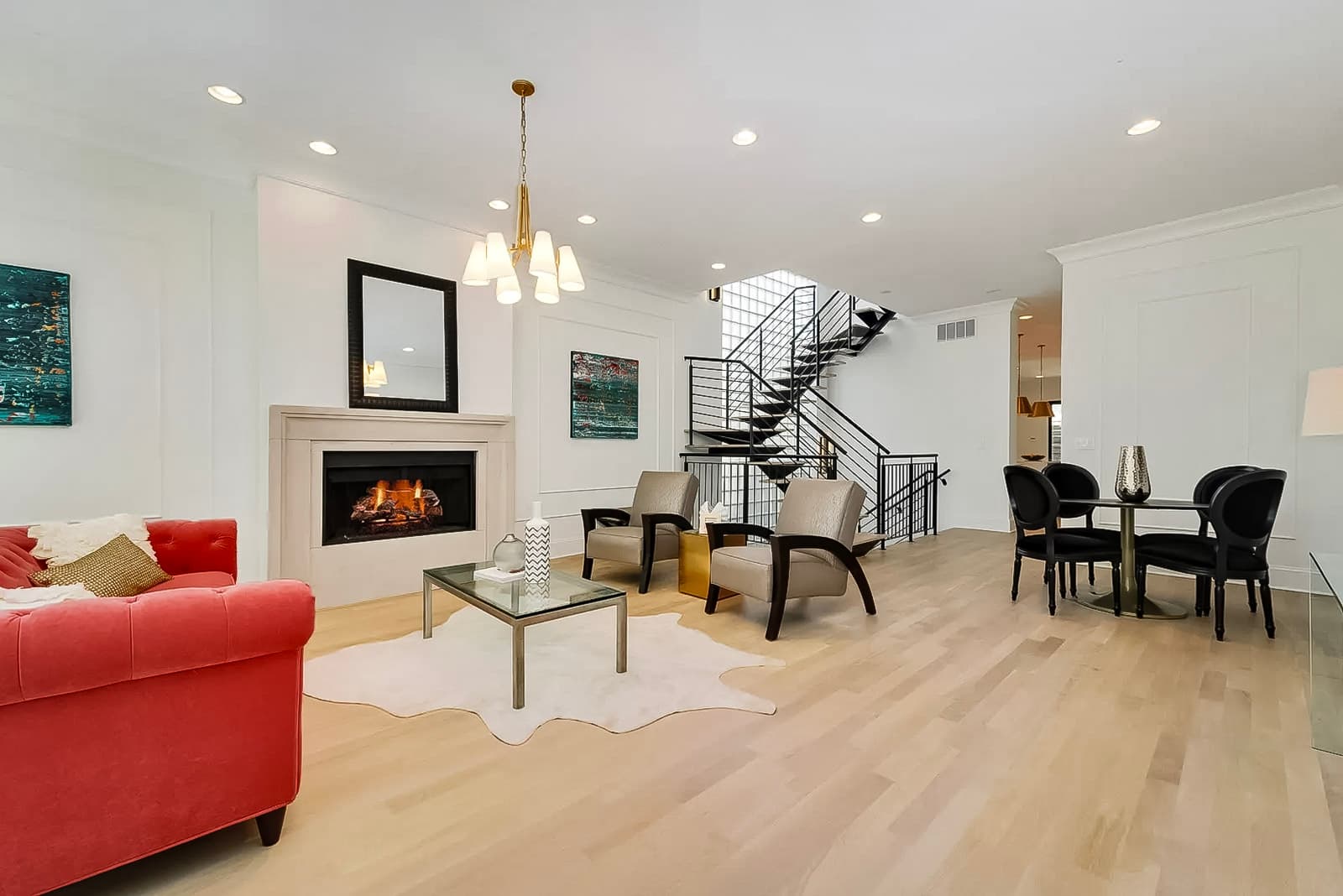 Modern living room with built-in fireplace, elegant red sofa, beige armchairs, black metal staircase, light wood flooring, and golden pendant lighting.